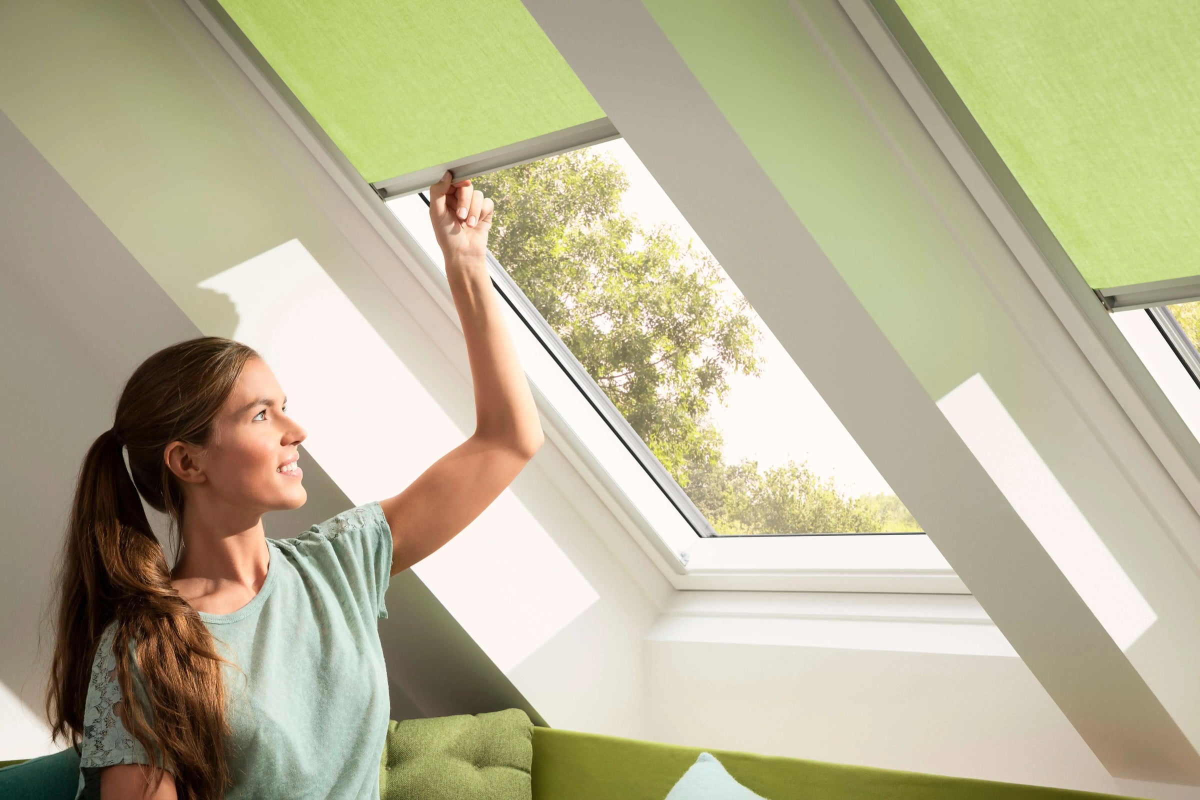 Woman adjusting green window blinds in a bright room with a view of trees.